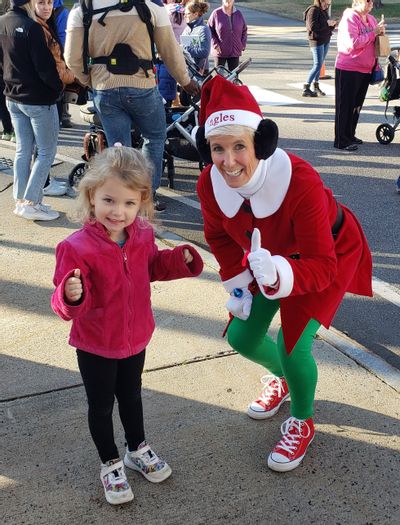 Photo is outdoors with a little girl standing next to a woman dressed as a holiday Elf in the foreground. The background has indistinguishable people on the sidewalk.