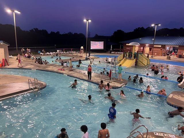 Families and children enjoy an evening of swimming and entertainment during Dive-In Movies at the Statesville Leisure Pool. Guests float in the illuminated pool and relax poolside while a movie plays on a large outdoor screen beside colorful water features and slides at dusk.