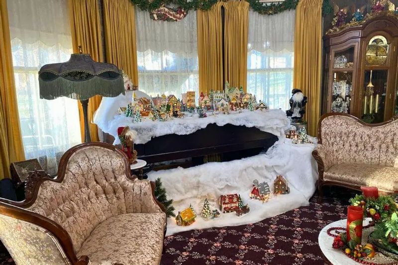 A holiday village display set atop a piano at Oakenwald Terrace during a Christmas open house, surrounded by Victorian-style seating, garlands, and festive décor.