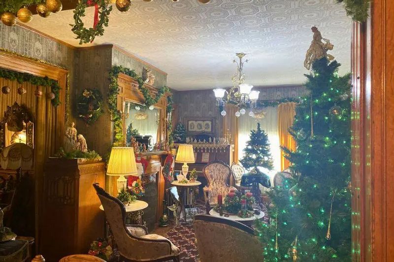 A cozy parlor at Oakenwald Terrace decorated for a Christmas open house, with lit Christmas trees, garlands, wreaths, and Victorian-style furnishings glowing in warm light.