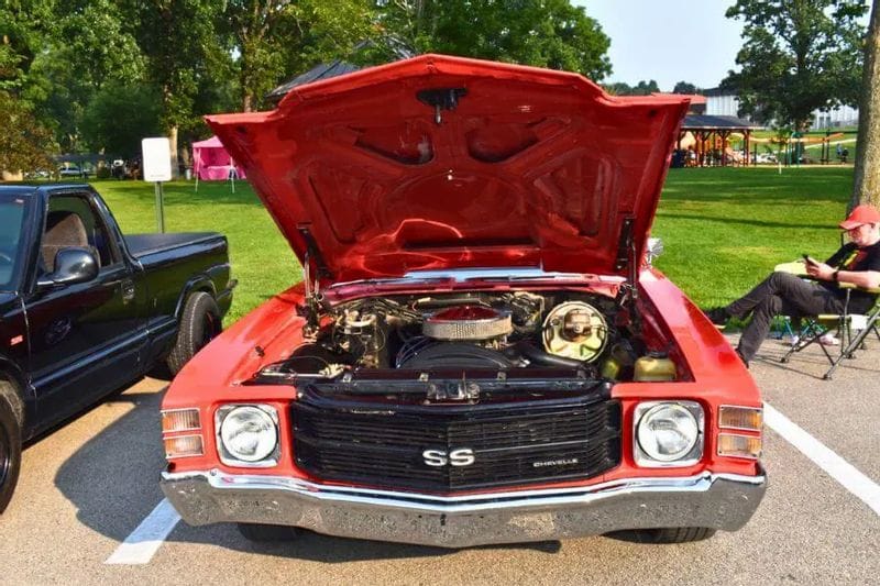 A red classic Chevrolet Chevelle SS with its hood open, showcasing the engine at a car show in Lanesboro, with green park space and spectators in the background.