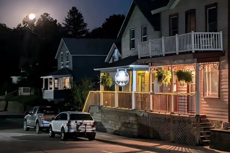 The Coffee Street Inn in Lanesboro illuminated at night, with porch lights glowing, hanging ferns along the railing, parked cars on the street, and a full moon rising above the trees.