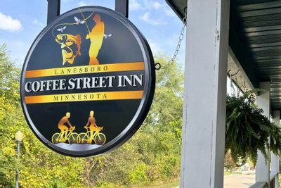 Hanging round sign for the Coffee Street Inn in Lanesboro, Minnesota, featuring illustrations of fishing and biking, mounted on a porch with trees in the background.