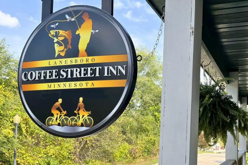 Hanging round sign for the Coffee Street Inn in Lanesboro, Minnesota, featuring illustrations of fishing and biking, mounted on a porch with trees in the background.