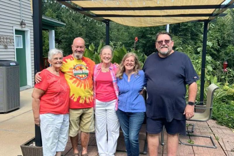 Five adults smiling and standing together on a shaded patio at the Coffee Street Inn in Lanesboro, surrounded by greenery and outdoor seating.