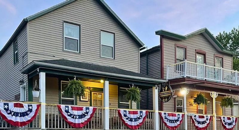 Exterior of the Coffee Street Inn in Lanesboro, Minnesota, showing two historic-style buildings with front porches decorated with patriotic bunting and hanging ferns in the evening light.