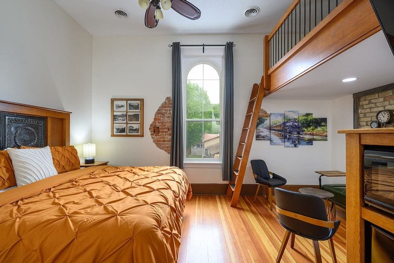 Spacious hotel room in Lanesboro featuring a king bed with warm-toned bedding, hardwood floors, a lofted sleeping area accessed by ladder, exposed brick accents, and a sitting area with chairs and artwork.