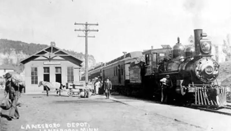 Historic black-and-white photograph of the Lanesboro railroad depot with a steam locomotive at the platform, people standing nearby, and horse-drawn wagons visible during the early railroad era in Lanesboro, Minnesota.