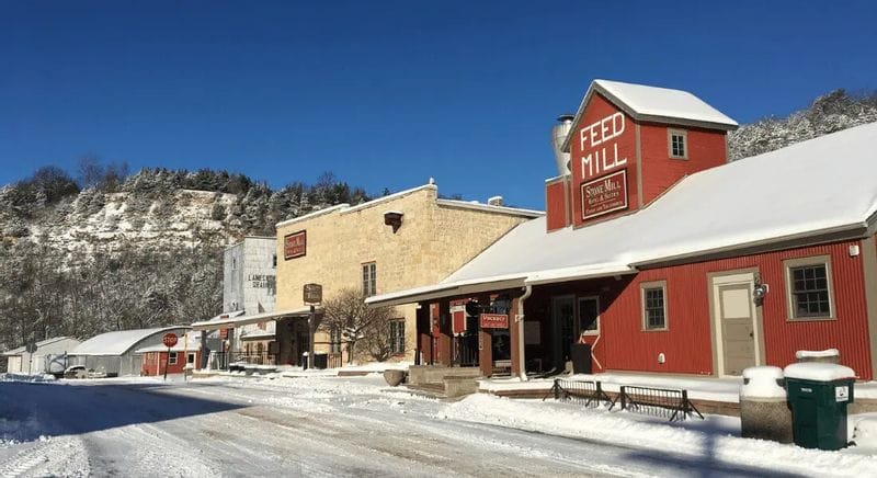 The Stone Mill Hotel & Suites and historic Feed Mill buildings line Parkway Avenue in Lanesboro during winter, with fresh snow covering rooftops and the street beneath clear blue skies and bluff views in the background.
