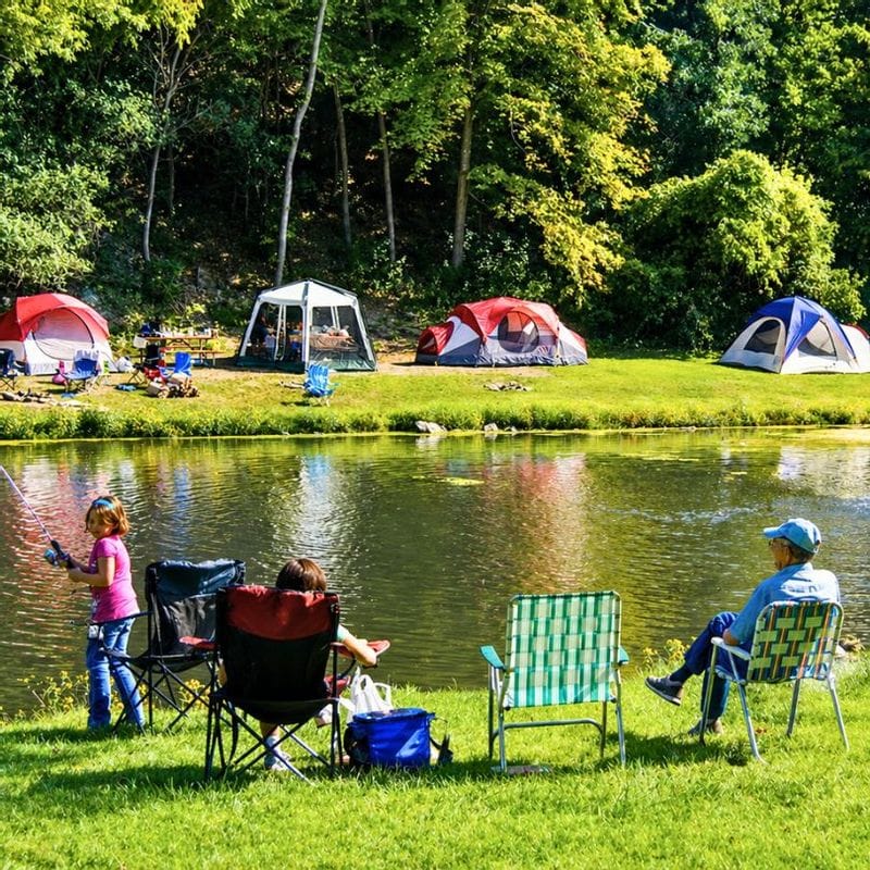 Family fishing beside a pond with tents and campers set up along a grassy riverside campground in Lanesboro, Minnesota, surrounded by trees in Bluff Country