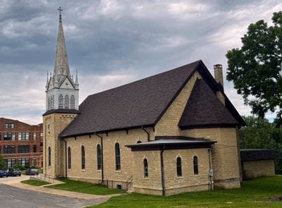 Historic stone church building in Minnesota with a tall steeple and cross, viewed from the street under a cloudy sky, representing local worship and faith communities