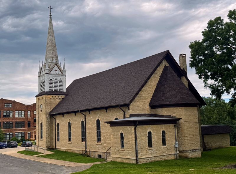 Historic stone church building in Minnesota with a tall steeple and cross, viewed from the street under a cloudy sky, representing local worship and faith communities