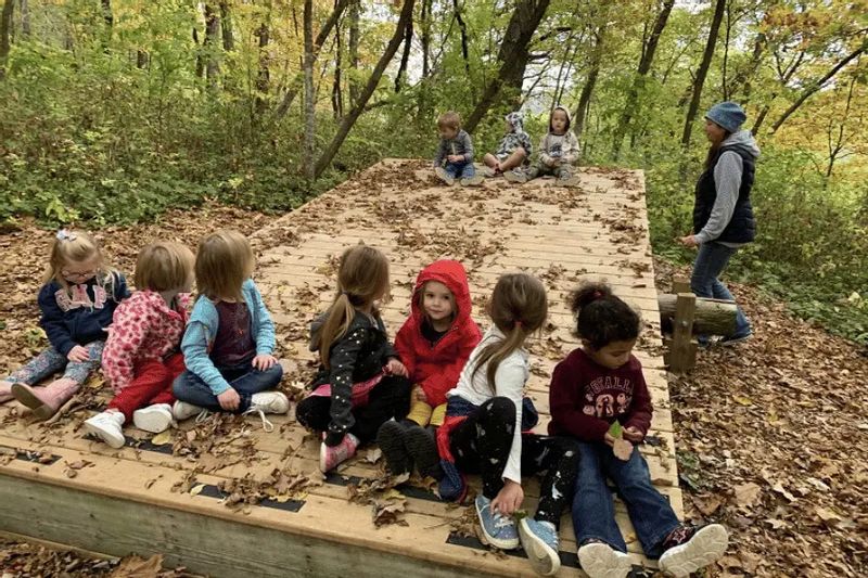 A group of preschool children sit together on a leaf-covered wooden platform in a forest while an educator stands nearby, part of an outdoor nature preschool activity surrounded by autumn trees.