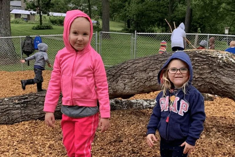 Two preschool children stand and smile near a fallen log in an outdoor play area, with other children exploring and climbing in the background at a nature-based preschool.
