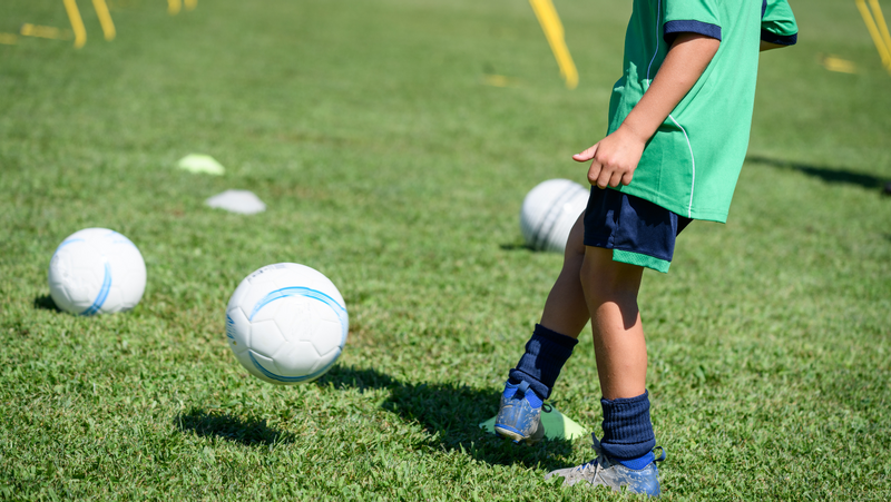 A child wearing a green soccer uniform practices dribbling a soccer ball across a grassy field during a sports camp activity. Several soccer balls and training cones are spread across the field in the background as part of the outdoor soccer drills.