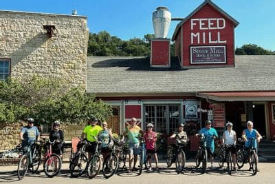 A group of cyclists pose with their bikes outside the Stone Mill Hotel & Suites and Feed Mill building in Lanesboro, gathering along Parkway Avenue before or after a ride on a sunny day.