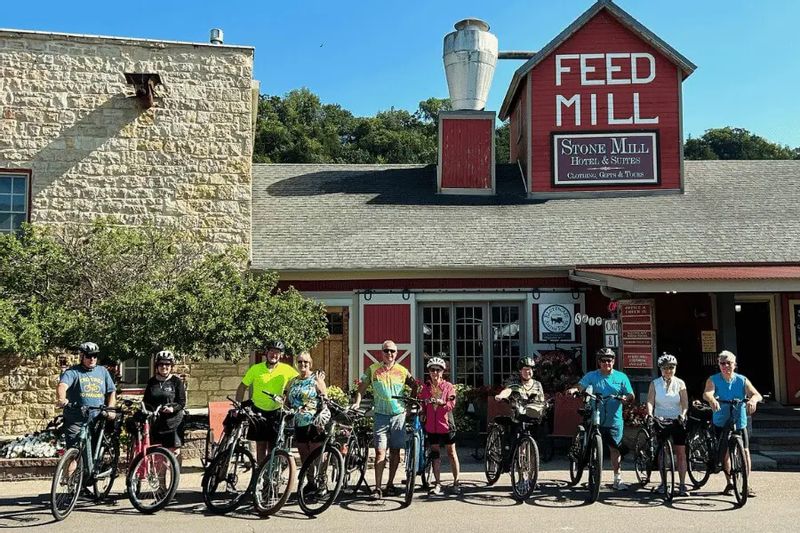 A group of cyclists pose with their bikes outside the Stone Mill Hotel & Suites and Feed Mill building in Lanesboro, gathering along Parkway Avenue before or after a ride on a sunny day.