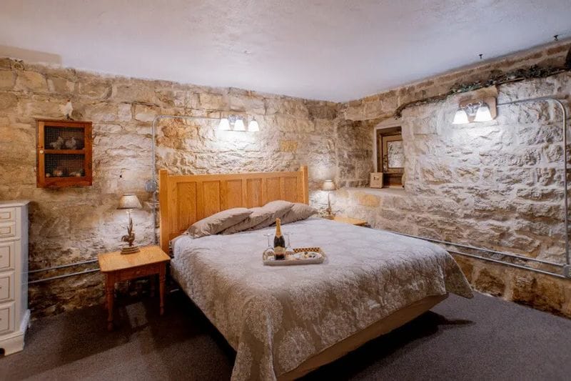 A cozy stone-walled guest room at the Stone Mill Hotel & Suites features a king bed with neutral bedding, warm lighting, rustic details, and a welcoming, historic ambiance.