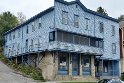 An aging blue wooden hotel building sits on a sloped street, featuring multiple stories, tall windows, and a covered porch supported by posts above a stone foundation, showing visible wear and weathering.