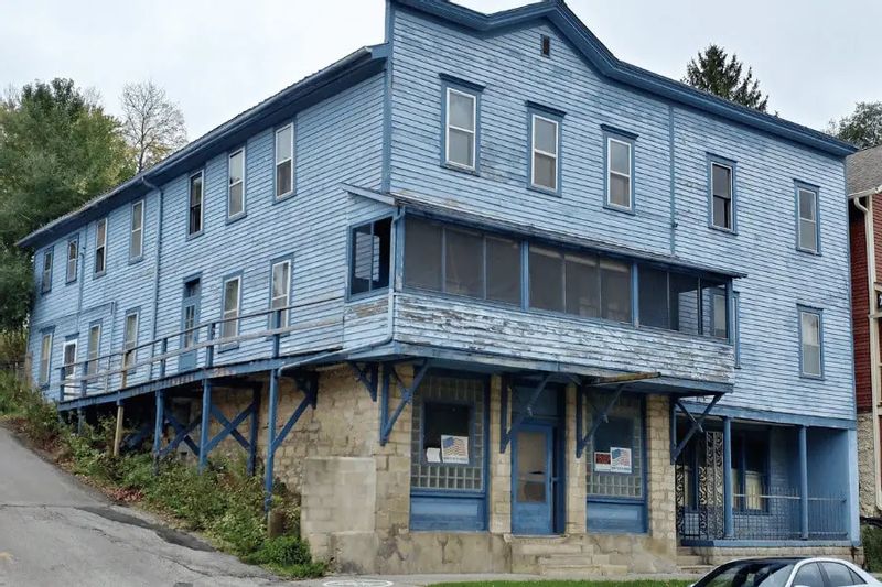 An aging blue wooden hotel building sits on a sloped street, featuring multiple stories, tall windows, and a covered porch supported by posts above a stone foundation, showing visible wear and weathering.