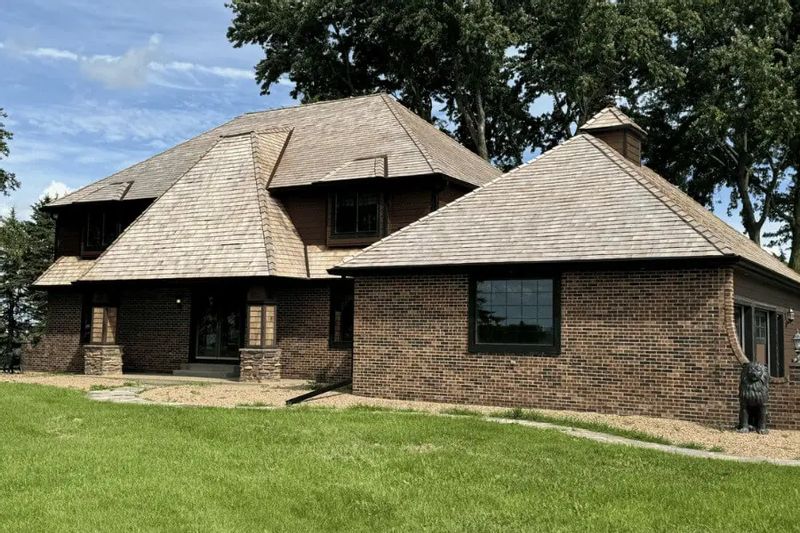 A well-maintained brick building with a steep, multi-angled roof covered in cedar shingles sits on a grassy lawn, with mature trees in the background under a clear summer sky.