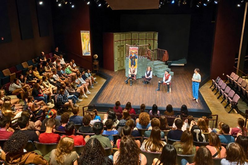 Audience members watch a Commonweal Theatre presentation from the seats surrounding the stage, with actors seated in front of a detailed theatrical set in Lanesboro.
