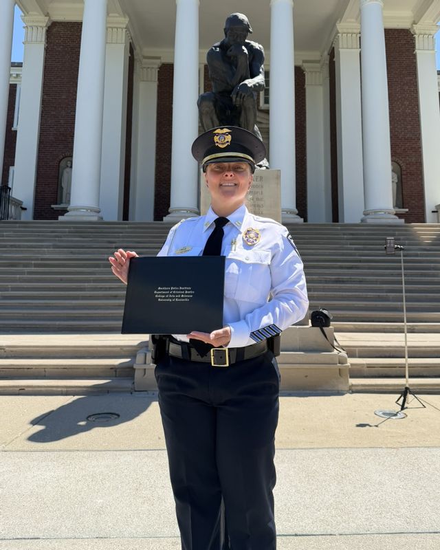 Captain Samantha Lindon of the Statesville Police Department stands outdoors in uniform while holding her graduation certificate from the Southern Police Institute leadership program. She poses on the steps of a large university building with white columns and a bronze statue in the background. Captain Lindon smiles toward the camera as she celebrates completing the prestigious leadership program.