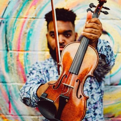 Musician Emanuel Wynter poses with a violin held toward the camera while standing in front of a brightly colored mural with swirling rainbow tones. He wears a patterned shirt and holds a violin bow in one hand while looking toward the viewer. The close-up composition highlights the violin and creates an artistic, vibrant atmosphere.