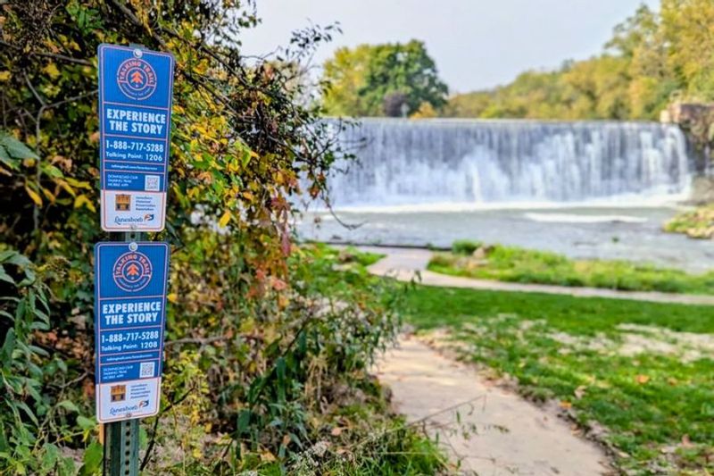 Talking Trail interpretive signs stand beside a path overlooking the Lanesboro Dam and Root River, surrounded by early fall foliage in Lanesboro, Minnesota.