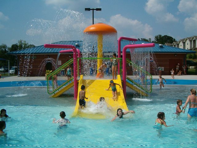 Children splash and play at the Statesville Leisure Pool on a sunny summer day. Kids slide down a bright yellow water slide beneath colorful water features spraying in every direction, while families enjoy the shallow pool area and interactive splash structures in the background.