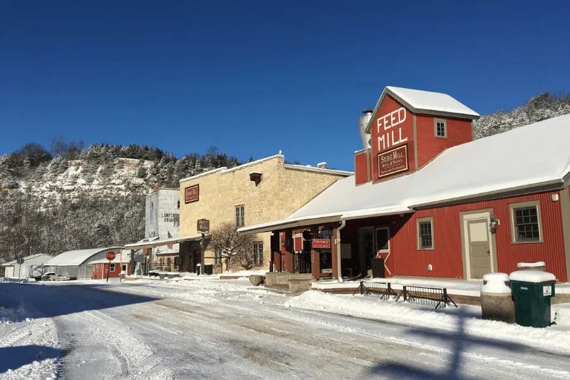 Snow-covered Stone Mill complex in Lanesboro, Minnesota, with the red Feed Mill building and historic storefronts lining a quiet winter street under a clear blue sky.