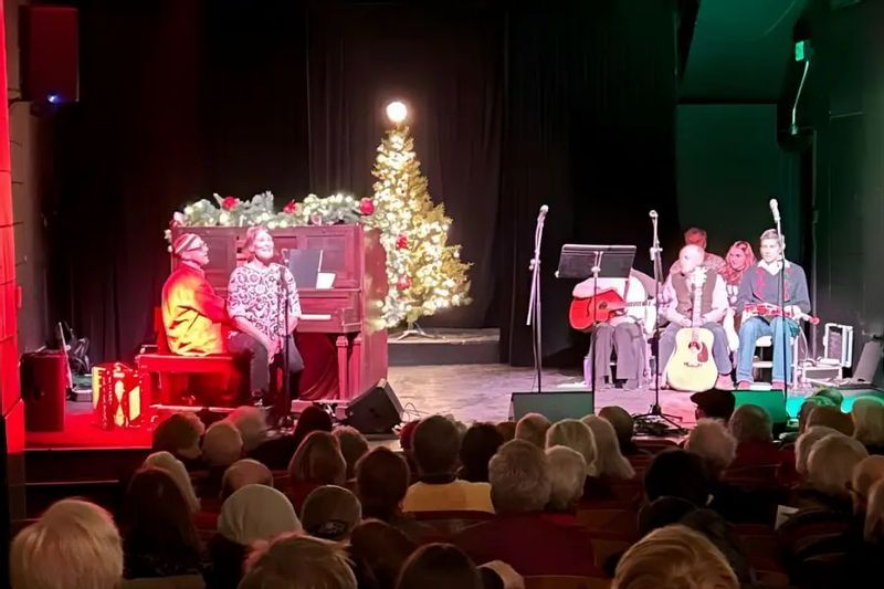 Holiday sing-along performance on a Lanesboro Arts stage, with musicians and singers seated under festive lighting, a decorated Christmas tree in the background, and a full audience watching from the theater seats.