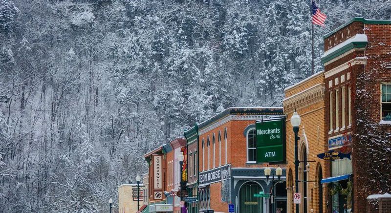 Snowy winter scene in downtown Lanesboro with historic brick storefronts, an American flag flying above the street, and snow-covered bluffs rising behind the buildings.