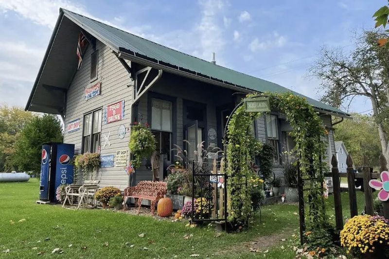 The Whalan Depot, a historic wooden building decorated with flowers and greenery, sits along a grassy lawn with an inviting front porch on a sunny day in Whalan, Minnesota.