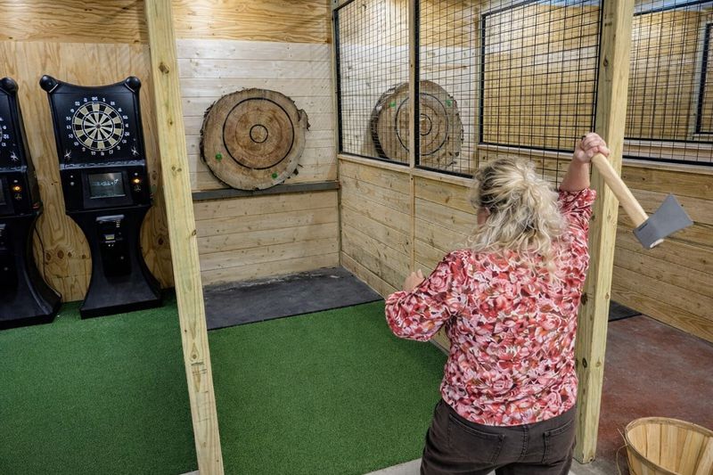 Woman prepares to throw an axe at a wooden target inside an indoor axe-throwing lane, with dartboards visible nearby and wood-paneled walls and green turf flooring surrounding the target area.