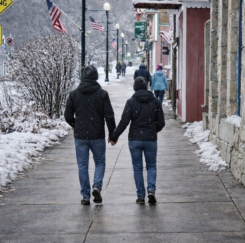 Couple holding hands while walking along a snowy downtown Lanesboro sidewalk lined with historic storefronts and American flags during winter.