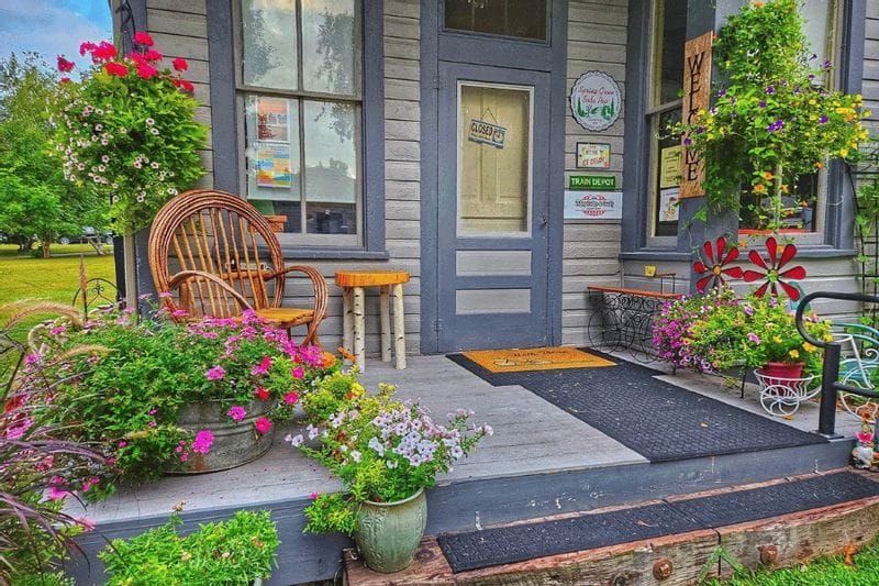 The front porch of the Whalan Depot is decorated with colorful flowers, potted plants, and vintage décor, creating a welcoming entrance to the historic shop in Whalan, Minnesota.