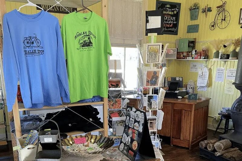 Inside the Whalan Depot gift shop, racks of branded T-shirts, postcards, stickers, and locally made items fill a cozy, colorful space celebrating the Root River Trail and small-town Whalan charm.