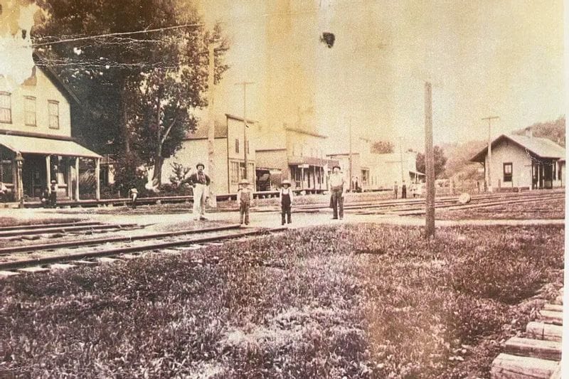 A historic sepia-toned photograph shows railroad tracks running through downtown Whalan, Minnesota, with early storefronts and residents standing near the tracks in the late 19th or early 20th century.