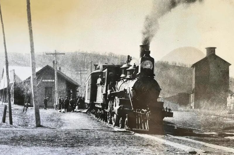 A historic black-and-white photograph shows a steam locomotive at the Whalan train depot, with smoke rising from the engine and people gathered along the tracks in early 20th-century Whalan, Minnesota.