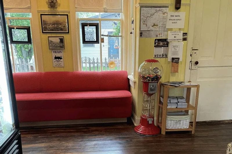 A cozy interior of the Whalan Depot features a red bench, vintage train photos, local maps, and a classic gumball machine, creating a welcoming small-town atmosphere for visitors.
