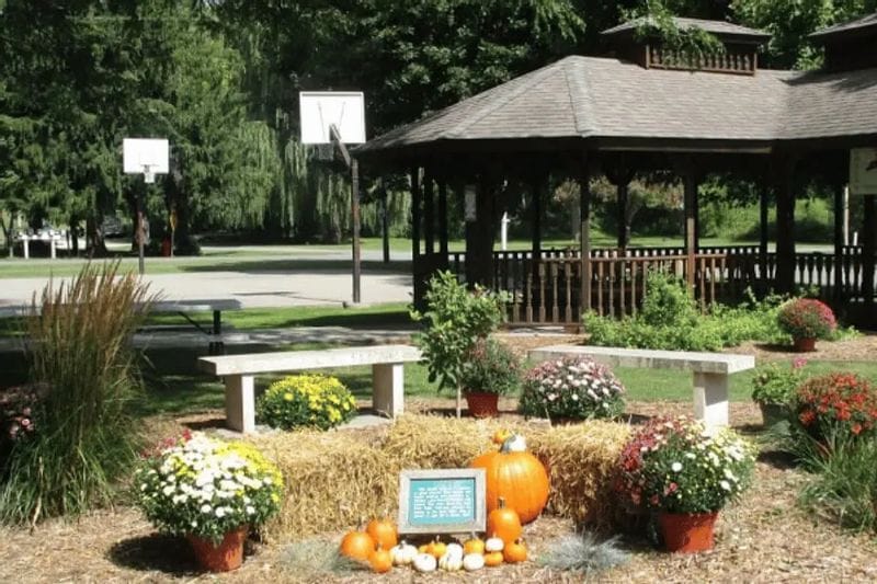 A gazebo in Whalan’s riverside park is decorated with pumpkins, hay bales, and fall flowers, with benches and a basketball court nearby on a sunny autumn day.