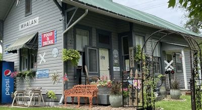 The Whalan Depot, a historic railroad-era building with colorful signs, flowers, and an “open” entrance, welcomes visitors to enjoy food, drinks, and small-town charm in Whalan, Minnesota.