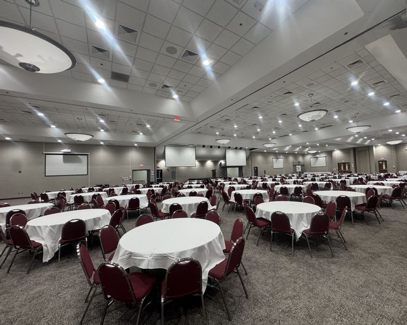 The Statesville Event Center is set up for a large gathering, with dozens of round tables covered in white tablecloths and surrounded by red chairs. The spacious ballroom features high ceilings, bright overhead lighting, multiple projection screens, and a stage area, creating a versatile venue for conferences, banquets, meetings, and community events.