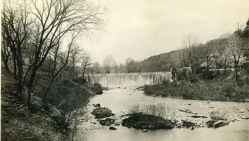 Historic black-and-white photograph of the Lanesboro dam spanning the Root River, with leafless trees and rocky riverbanks during the town’s early years.
