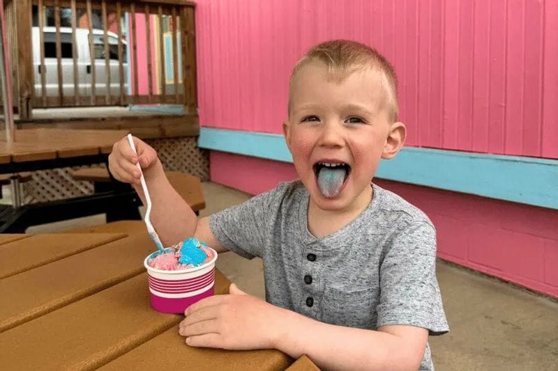 Young child sitting at an outdoor table, smiling with blue tongue while eating colorful ice cream from a cup outside a bright pink ice cream shop in Lanesboro, Minnesota.