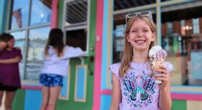 Smiling child holding a scoop of ice cream in front of a brightly colored ice cream shop in downtown Lanesboro, Minnesota, with other kids waiting in the background.