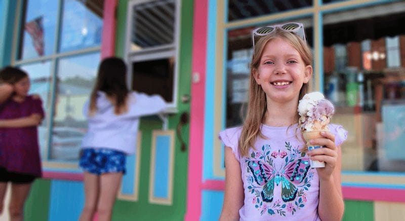 Smiling child holding a scoop of ice cream in front of a brightly colored ice cream shop in downtown Lanesboro, Minnesota, with other kids waiting in the background.