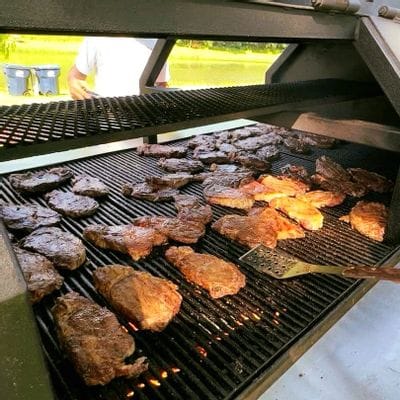 Multiple steaks grilling over open flames on a large outdoor barbecue grill during a community steak fry in Lanesboro, with a spatula resting on the grate and a person standing nearby.