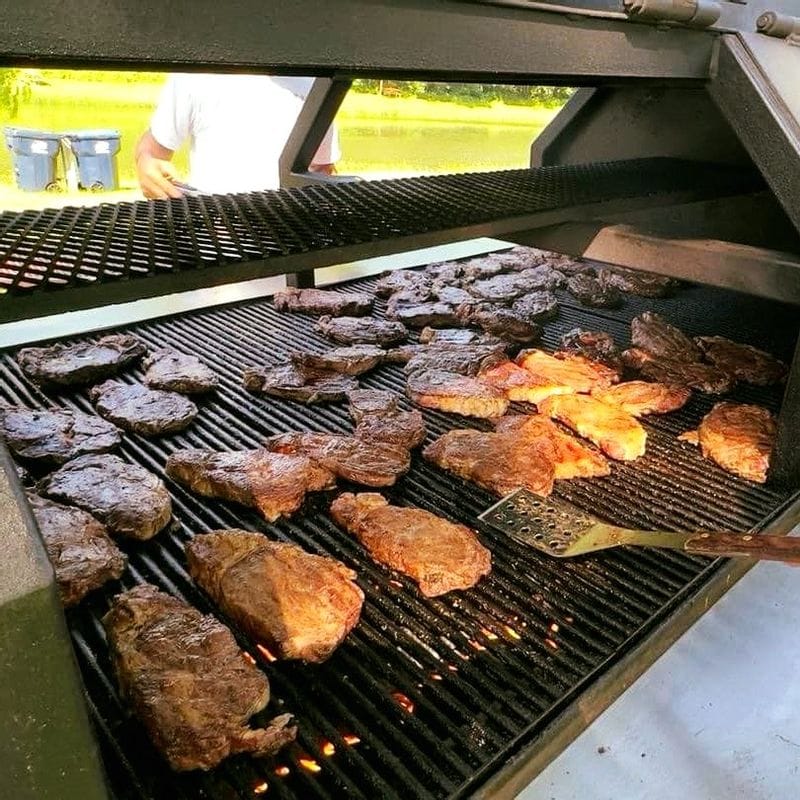 Multiple steaks grilling over open flames on a large outdoor barbecue grill during a community steak fry in Lanesboro, with a spatula resting on the grate and a person standing nearby.
